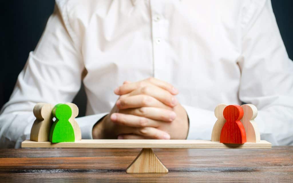 A person in a white shirt sits at a table with hands clasped, embodying the role of a divorce mediator. In front, a balance scale holds three green wooden figures on one side and three red on the other, symbolizing negotiation and equitable decision-making.