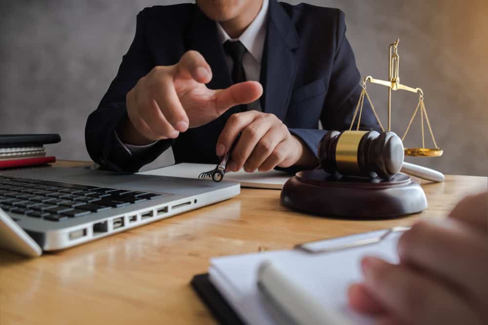 A person in a suit gestures while sitting at a desk with a laptop, notepad, gavel, and scales of justice. Another hand is seen in the foreground, holding a pen over a clipboard.