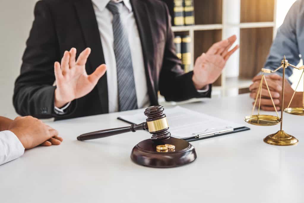 A divorce mediator in a suit sits at a desk with paperwork, gesturing with hands raised. A gavel, two rings, and a balance scale on the table signal the solemnity of the mediation process. Two individuals are partially visible, facing the mediator in this legal setting.