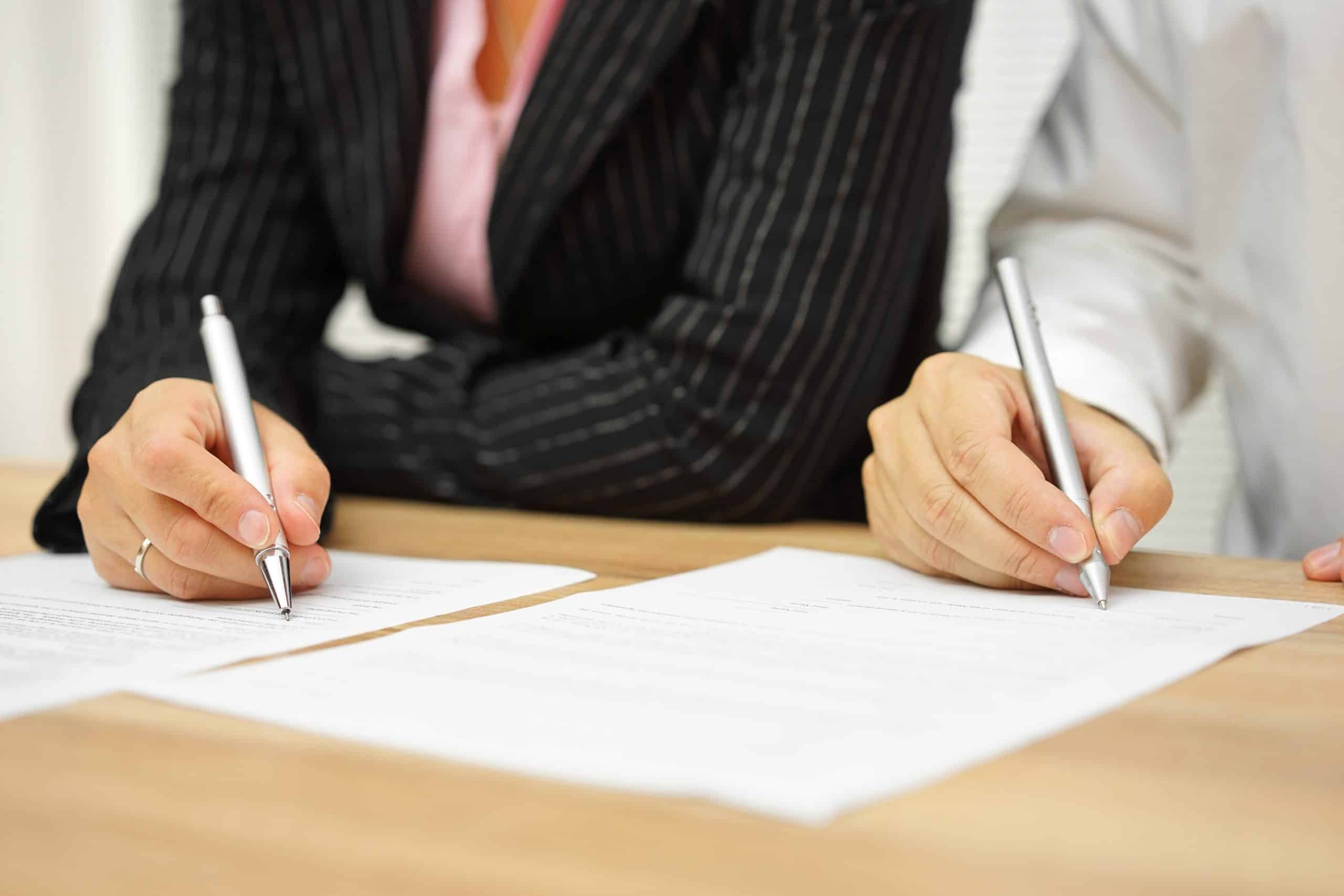 Two people sit side by side at a table, both dressed in formal attire. They are holding pens and writing on separate sheets of paper placed in front of them. The presence of a divorce mediator subtly shifts the focus to their hands as they carefully navigate the documents.