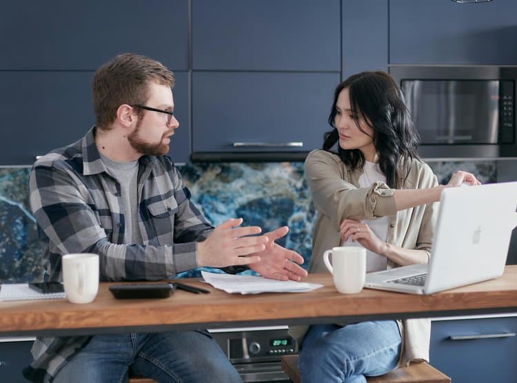 A man and woman sit at a kitchen counter with a laptop, coffee mugs, and papers. The man gestures while speaking, and the woman points at the laptop screen. They appear to be having a discussion.