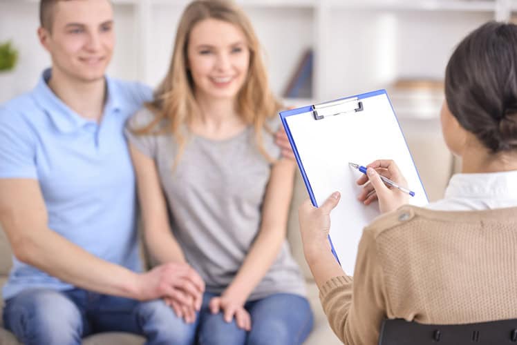 A couple sits closely together, smiling, while a person with a clipboard and pen, seen from behind, appears to be consulting or counseling them in a bright, comfortable room.