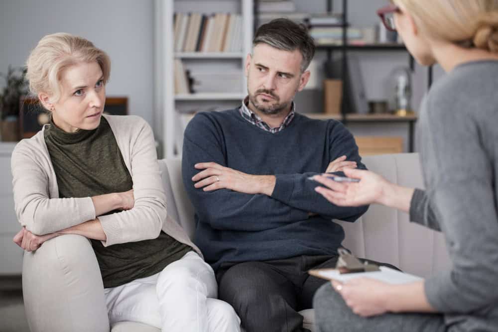A woman and man sit on a couch with arms crossed, looking serious, while another person, partially visible, speaks to them and holds a clipboard, suggesting a therapy or counseling session.