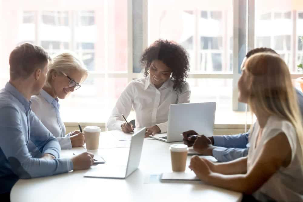Five colleagues sit around a table with laptops and coffee cups, engaging in a discussion and taking notes in a bright office setting.
