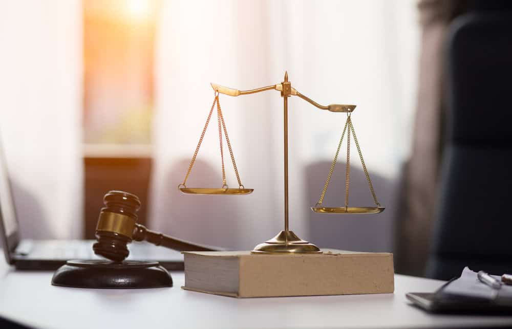 A brass balance scale and a wooden judge’s gavel rest on a desk with a closed book, symbolizing justice and law, with sunlight streaming through sheer curtains in the background.
