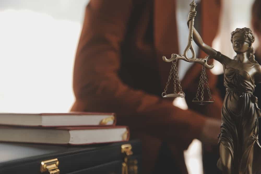 A bronze statue of Lady Justice holding scales stands on a desk with a stack of books. A person in a brown blazer is partially visible in the background.