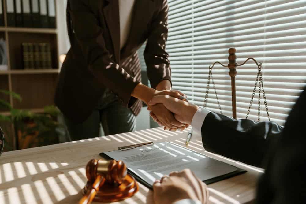 Two individuals are shaking hands across a desk in an office, with sunlight streaming through window blinds. A contract, a gavel, and scales of justice are on the desk, suggesting a legal or business agreement.