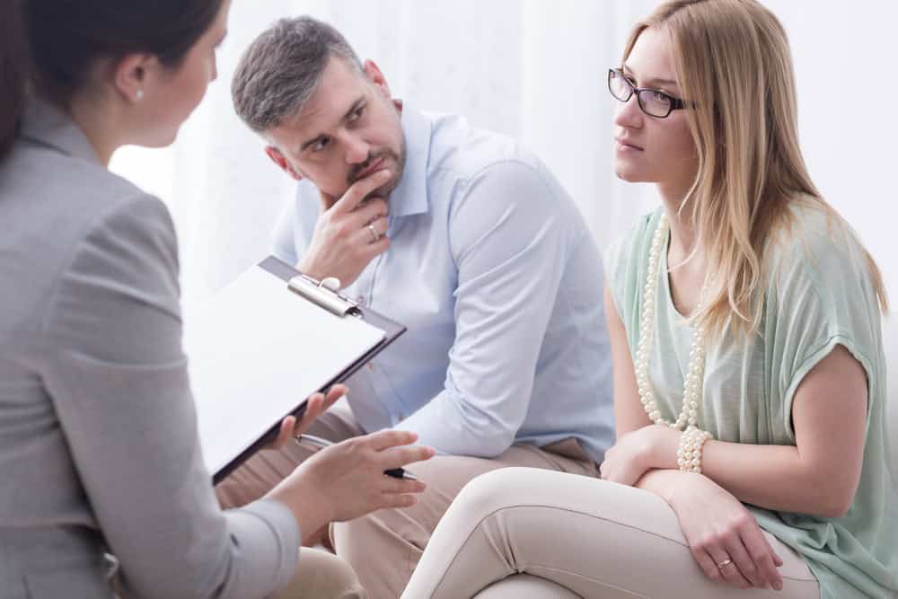 A couple sits together on a couch, listening attentively to a therapist holding a clipboard during a counseling session. The woman looks thoughtful, while the man rests his chin on his hand.