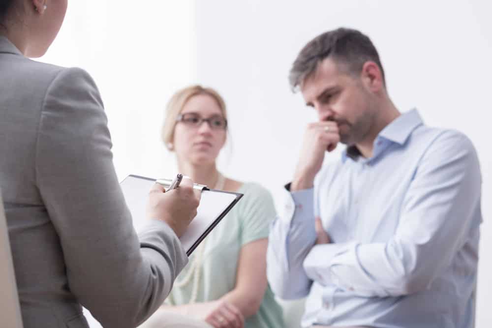 A man and woman sit together, looking serious, while a person in a suit holds a clipboard and pen, possibly conducting a counseling or therapy session.