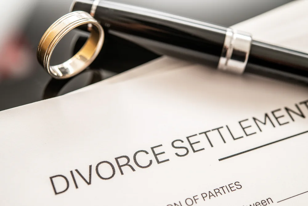 A gold wedding ring and a black pen rest on a document titled "Divorce Settlement," indicating the signing of a legal agreement related to divorce.